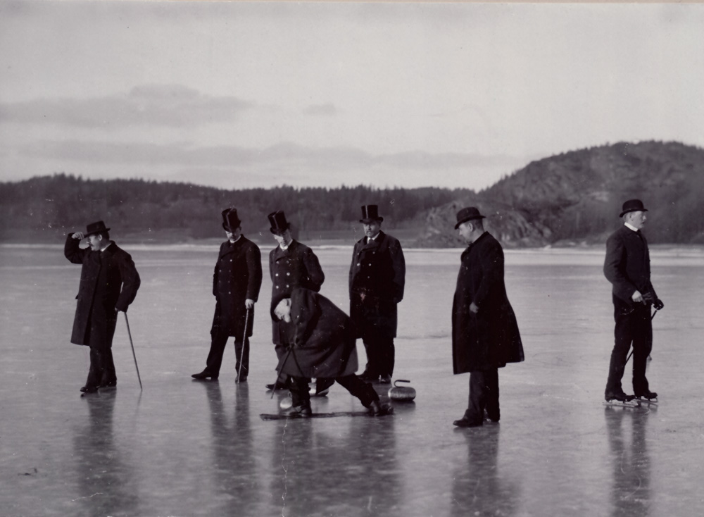 7 välklädda herrar spelar curling på havsis. En har skridskor på fötterna. Bild från 1887