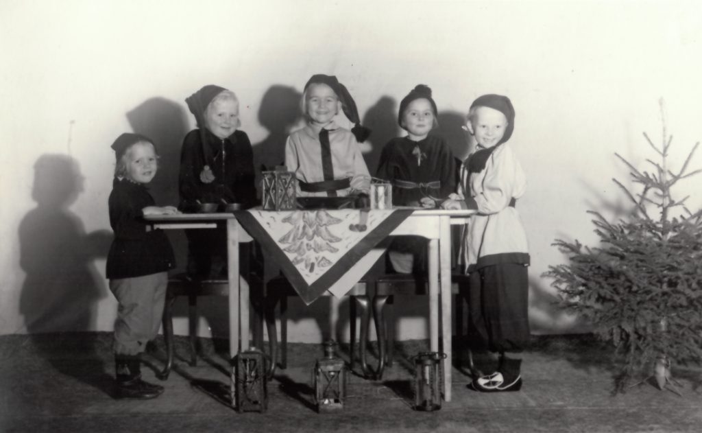Äldre svartvitt foto föreställande fem barn utklädda till tomtar som står runt ett bord.