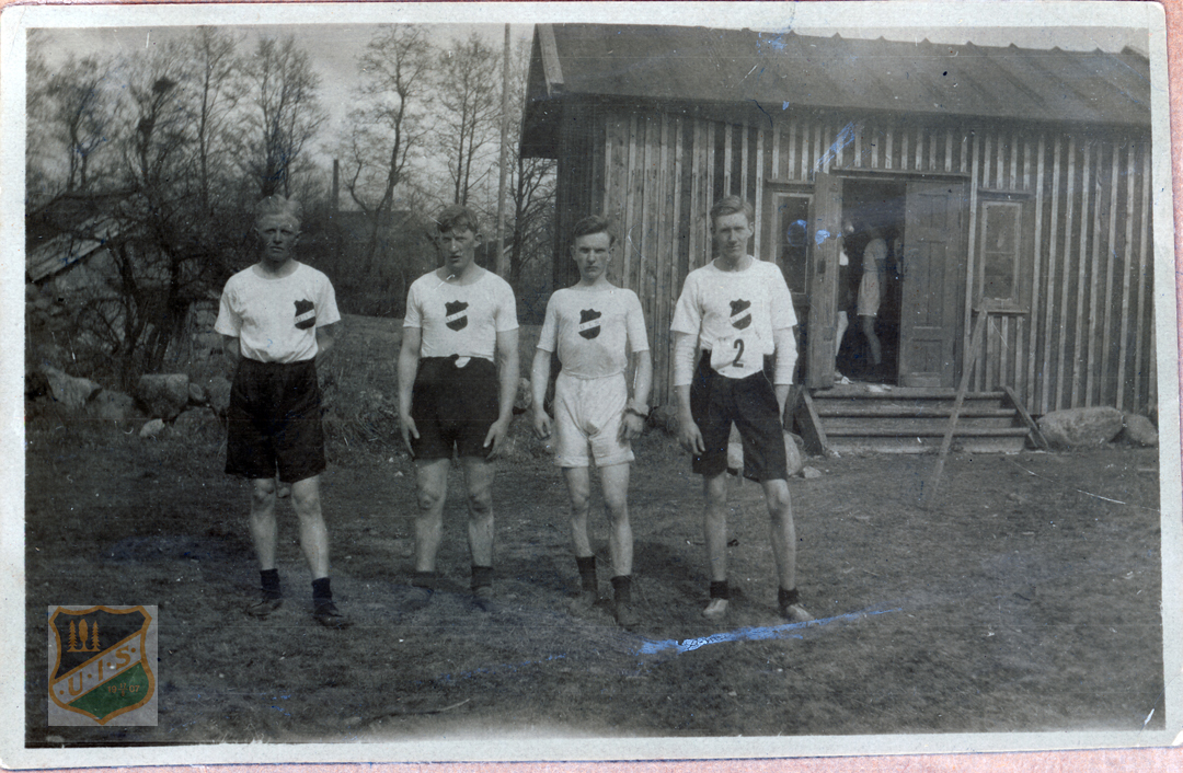Fyra löpare uppställda framför mindre byggnad. Löparna är tidsenligt klädda för året 1922.
