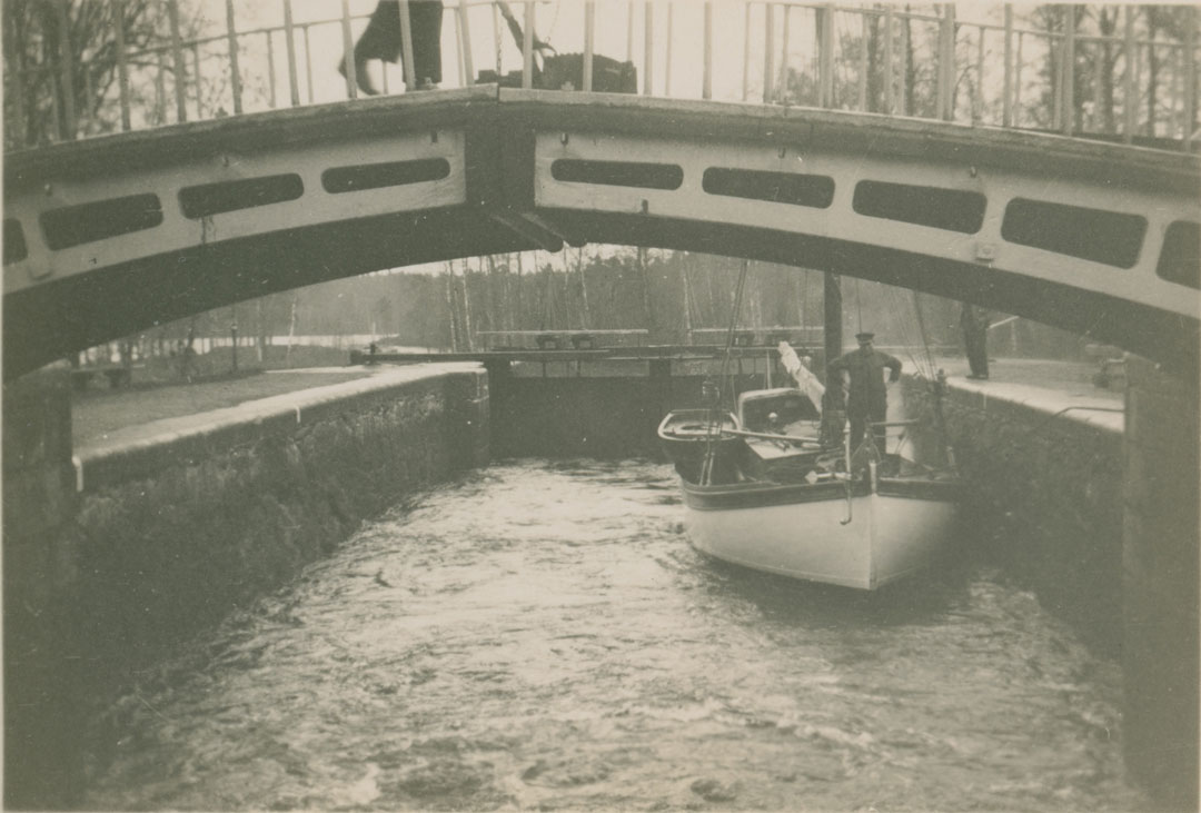 Seger II slussas på väg hemåt. En person står i fören på båten och en på sidan av slussen. I förgrunden någon form av bros som person går på.