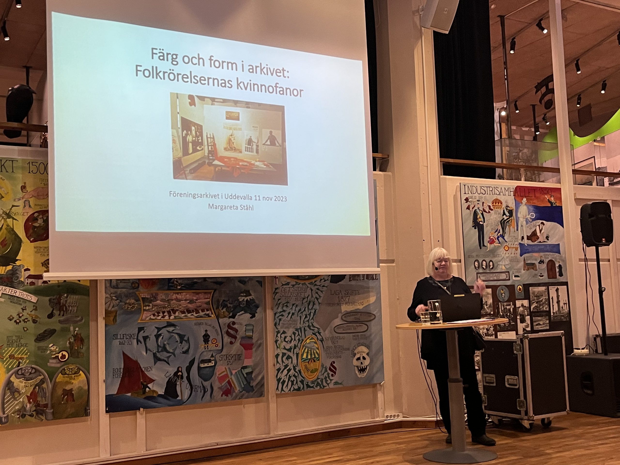 Närbild av föredragshållaren och en "slide" på duken. Överst texten Färg och form i arkivet: Folkrörelsernas kvinnofanor. där under otydlig överblicksbild. Under bilden finns texten Föreningsarkivet i Uddevalla 11 nov 2023 Margareta Ståhl